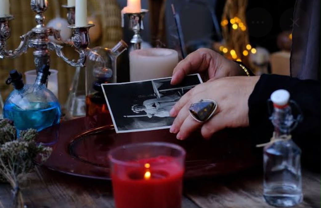 Mâini ținând o fotografie lângă o lumânare roșie și recipiente de ritual